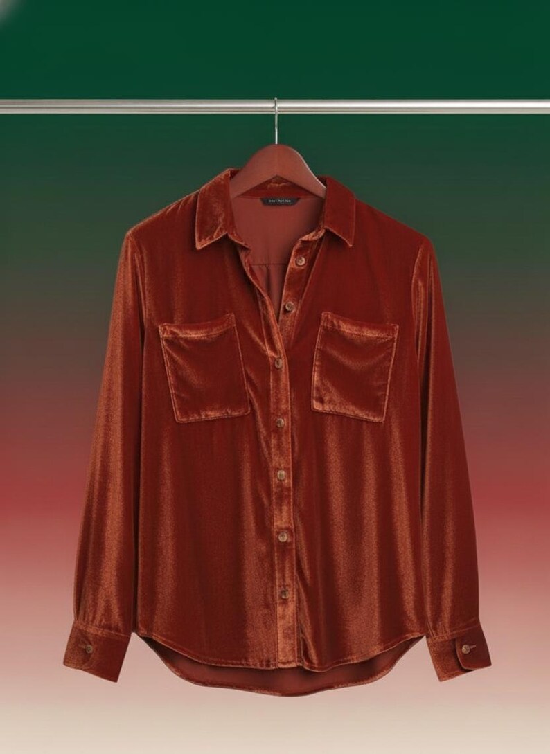 May include: A burnt orange velvet shirt with long sleeves, a collared neckline, and two front pockets. The shirt is button-down and hangs on a wooden hanger against a green and red gradient background.