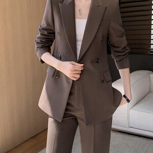 Pode incluir: Um fato de duas pe&ccedil;as castanho com um blazer de abotoamento duplo e cal&ccedil;as a condizer. O blazer tem lapelas entalhadas e detalhes de bot&otilde;es. O fato &eacute; de cor lisa, adequado para neg&oacute;cios ou eventos formais.