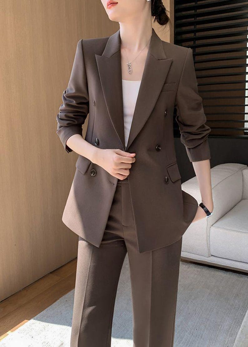 Pode incluir: Um fato de duas pe&ccedil;as castanho com um blazer de abotoamento duplo e cal&ccedil;as a condizer. O blazer tem lapelas entalhadas e detalhes de bot&otilde;es. O fato &eacute; de cor lisa, adequado para neg&oacute;cios ou eventos formais.