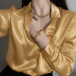Pode incluir: Uma blusa de cetim amarelo dourado com gola e mangas compridas. Um colar e uma pulseira de corrente dourada são visíveis. A blusa é abotoada na frente e combinada com calças pretas.