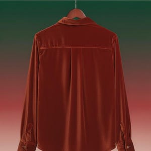 May include: A rust-coloured velvet shirt with long sleeves, a collared neckline, and buttoned cuffs. The back of the shirt features a vertical pleat detail. The shirt is hanging on a wooden hanger against a gradient background.