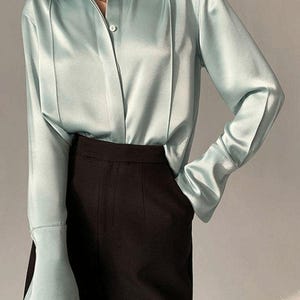 Könnte beinhalten: Eine hellblaue Satinbluse mit klassischem Kragen und langen Ärmeln. Die Bluse hat eine Knopfleiste und wird mit einer schwarzen Hose kombiniert. Die Ärmel haben ein ausgestelltes Design an den Manschetten. Eine goldene Halskette ist sichtbar.