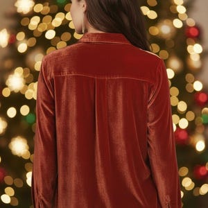 May include: A burnt orange velvet shirt with long sleeves and a button-down design. The shirt has a traditional collar and a fitted shape. The background shows a blurred Christmas tree with festive lights, suggesting a holiday theme.