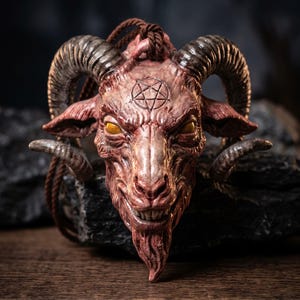 May include: A pendant shaped like a goat head with large curved horns, yellow eyes, and a visible pentagram symbol on the forehead. The piece features a textured, reddish-brown finish and is suspended from a dark brown braided cord necklace.