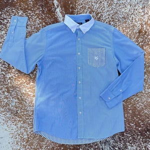 May include: Light blue button-down shirt with a white collar and a pocket featuring vertical blue and white stripes. The shirt has long sleeves and a striped pattern on the side and bottom. A classic style.