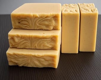 Può includere: Diverse saponette artigianali nei toni del giallo e beige. Tre saponette sono impilate, con un motivo a spirale decorativo sulla superficie superiore. Due saponette aggiuntive sono in piedi, con un design intagliato simile.