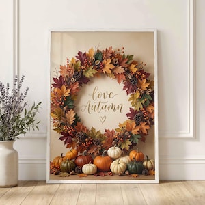 May include: A framed wall art print featuring a circular wreath made of multicoloured autumn leaves, pinecones, and berries surrounding the text love Autumn with a small heart. Below the wreath are several pumpkins in shades of orange, white, and green.