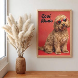 May include: A framed print with a brown dog wearing heart-shaped sunglasses on a red background. The print says "Cool Dude" in black script. A wooden vase with dried pampas grass is to the left of the print.