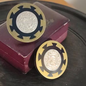 May include: Pair of vintage gold-toned cufflinks with a circular design. Each cufflink features a silver-colored center with an embossed pattern, surrounded by a black and gold border. The cufflinks are displayed on a burgundy box.