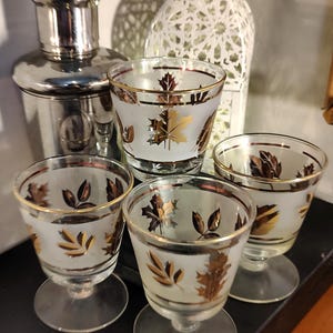 May include: Four vintage frosted glass cordial glasses with gold leaf designs. The glasses have a clear stem and a gold rim. A silver cocktail shaker is in the background.