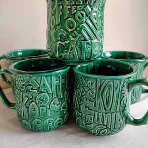May include: A set of four emerald green ceramic mugs featuring a raised, embossed texture with the word coffee written in various languages including Kawa, Kaffee, and Kopi. Each mug has a glossy finish, a sturdy handle, and a cylindrical shape.