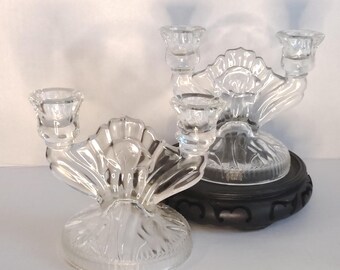 May include: Two clear glass candle holders with a shell-like design. Each holder has three candle cups. One holder sits on a black, round base. The other holder is on a clear, round base. The candle holders are decorative.