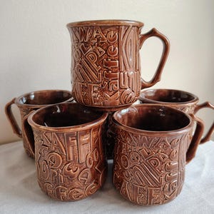 May include: A set of five brown ceramic mugs with a glossy finish, each featuring an embossed, repeating pattern of the word coffee in various languages and scripts, including the word Kaffee clearly visible on the top center mug.