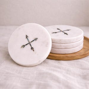 May include: White marble coasters with an inlaid arrow design. The coasters are round and stacked on a wooden tray. The arrow design is black and iridescent. The coasters are on a white linen surface.