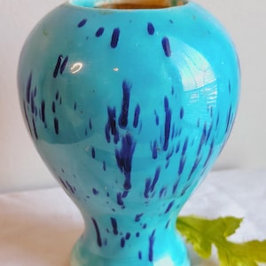 May include: A turquoise ceramic vase with a rounded body and a flared base. The vase is adorned with dark blue splatter patterns. The interior of the vase is a contrasting brown color. A decorative piece for home decor.