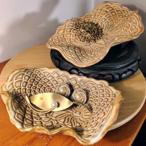 May include: Two decorative ceramic leaf-shaped dishes with textured patterns. One dish holds a gold-toned brooch, while the other contains a gold-toned leaf-shaped pin and a pair of pearl earrings. The dishes are on a wooden surface.