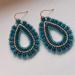May include: A pair of teardrop-shaped dangle earrings featuring dark teal faceted beads and a central inner border of small, light-colored seed beads. The earrings are constructed with gold-toned metal hooks for attachment.
