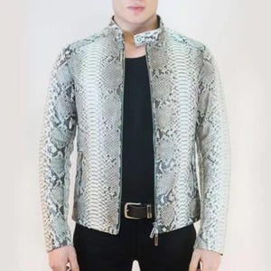 Op de afbeelding: Een jas met slangenpatroon in grijstinten en wit. De jas heeft een opstaande kraag, een ritssluiting aan de voorkant en lange mouwen. Het model draagt een zwart shirt en een zwarte broek met een bruine riem.