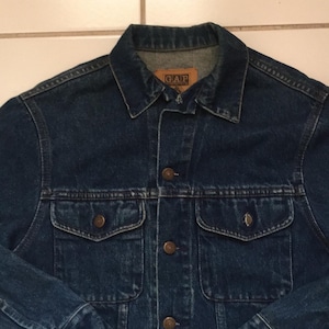 May include: A dark blue denim jacket with a classic collar, button fastenings, and two chest pockets. The jacket features the "GAP" logo on a small tag at the collar. The jacket is a vintage style.