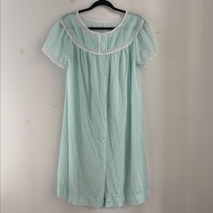 May include: A light blue vintage nightgown with short sleeves and a button-down front. The nightgown features a white lace trim around the neckline and sleeves. The garment is hanging on a black hanger.