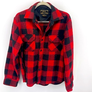 Puede incluir: Camisa de franela a cuadros rojos y negros con cierre de botones y mangas largas. La camisa tiene dos bolsillos en el pecho y una etiqueta que dice "Rugged Wear" con un logotipo de montaña.