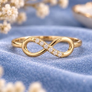 May include: A gold ring featuring an infinity symbol centrepiece. One side of the infinity loop is set with five small, clear, round-cut stones that sparkle against the polished gold band. The design is continuous, smooth, and elegant in its form.