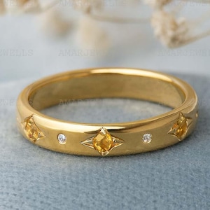 Pode incluir: Um anel dourado com elementos em forma de estrela contendo pedras preciosas amarelas e pedras transparentes. O anel é apresentado sobre um fundo azul claro. O design é simétrico.