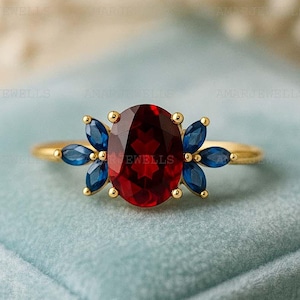 May include: A gold ring with a large, oval-cut, deep red gemstone at its centre. The ring is adorned with blue gemstones on either side, arranged in a floral pattern. The ring is presented on a light blue surface.