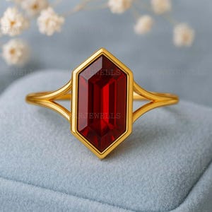 May include: A gold ring featuring a large, rectangular, faceted, deep red gemstone set in a hexagonal bezel. The ring's band splits into two delicate gold strands on either side of the gemstone, creating an open design.