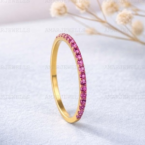 Op de afbeelding: Een gouden ring met een band van kleine, helder roze edelstenen. De ring staat tegen een zachte, witte achtergrond met delicate, gedroogde bloemen op de achtergrond. De ring is een delicaat sieraad.
