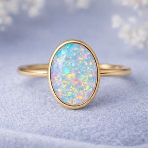 May include: A gold ring featuring an oval opal gemstone. The opal has a shimmering, iridescent quality with hues of blue, yellow, and green. The ring's band is a simple, polished gold.