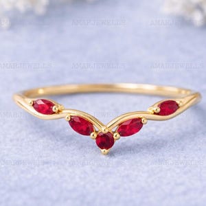 Op de afbeelding: Een goudkleurige ring met een gebogen ontwerp, bezet met vijf ovale, levendige rode edelstenen. De ring wordt gepresenteerd op een lichtblauwe ondergrond, die de elegante vorm van het sieraad benadrukt. De merknaam "AMARJEWELLS" is zichtbaar.