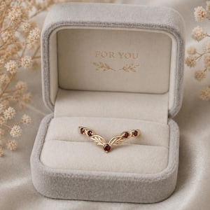May include: A gold-colored ring with a leaf design and small red gemstones, presented in a gray velvet box. The box is open, revealing the ring and the words "FOR YOU" inside. The ring is a V-shape.