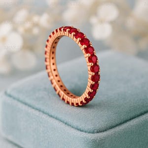 May include: A rose gold eternity ring featuring a continuous row of vibrant red gemstones. The ring is set against a soft blue background, highlighting the color and sparkle of the gemstones. The ring is a classic design.