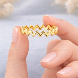 May include: A gold-colored ring with a zigzag design. The ring is held between two fingers, showcasing its intricate pattern. The ring's design is a series of connected chevrons, creating a unique and eye-catching look. The ring is a piece of jewelry.