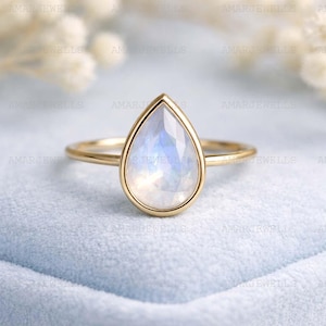 Pode incluir: Um anel de ouro com uma pedra da lua iridescente em forma de pêra. A gema está engastada em uma moldura de ouro e assenta-se sobre uma fina banda de ouro. O anel é exibido em uma superfície azul clara, com a marca "AMARJEWELLS" visível.