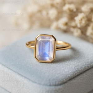 Könnte beinhalten: Ein goldener Ring mit einem rechteckigen, irisierenden Edelstein. Der Edelstein hat eine hellblaue und weiße Farbe. Der Ring liegt auf einer hellblauen Samtoberfläche. Ein Schmuckstück.