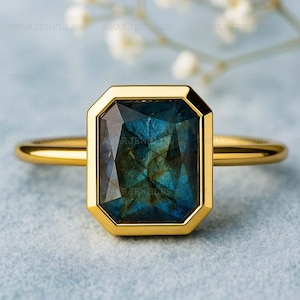 Puede incluir: Un anillo de oro con una piedra preciosa rectangular, iridiscente, azul-verdosa. La piedra preciosa está engastada en un bisel de oro, y la banda es de oro liso. El anillo se muestra sobre una superficie azul claro.