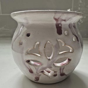 Może przedstawiać: Biały ceramiczny dyfuzor olejków eterycznych z kwiatowym wycięciem. Dyfuzor ma zaokrąglony kształt z rozszerzoną krawędzią i małymi otworami. Bordowe smugi dodają dekoracyjnego charakteru na zewnątrz.