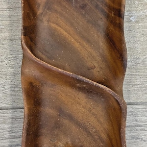 Puede incluir: Una bandeja de servir de madera hecha a mano con un diseño único y fluido. La bandeja es de madera marrón oscuro y presenta una forma orgánica tallada con un canal central curvo. La veta de la madera es visible.