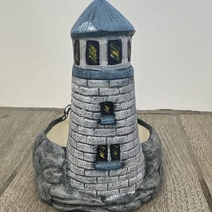 May include: A ceramic lighthouse planter with a blue roof and grey brickwork. The lighthouse has three windows with yellow and black details. The base is a grey rock formation, and the planter is empty.