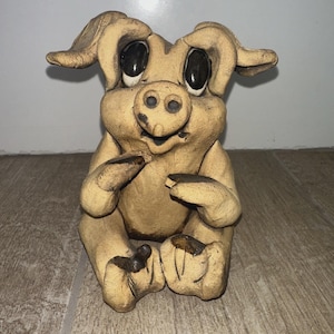 May include: A tan ceramic pig figurine with large black eyes, sitting with its legs crossed. The pig has large ears and a smiling expression. The figurine is handmade and has a rustic appearance.