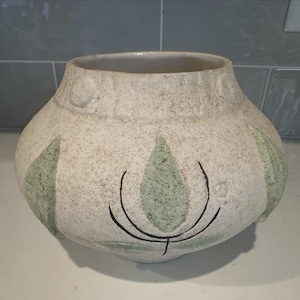 Puede incluir: Un jarrón de cerámica moteada de color blanquecino, con un cuerpo ancho y un cuello estrecho. El jarrón presenta diseños de hojas verdes con detalles de líneas negras. El jarrón tiene una superficie texturizada y una paleta de colores neutros, adecuada para la decoración del hogar.