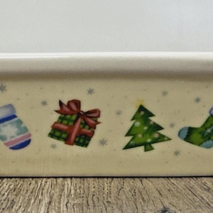 May include: Cream-coloured rectangular ceramic dish with a festive Christmas design. The design includes a mitten, a wrapped gift, a Christmas tree, and a stocking. The dish has a white rim and is decorated with small blue stars.