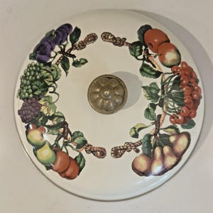 Könnte beinhalten: Ein runder, cremefarbener Keramikdeckel mit einem dekorativen Obst- und Rankenmuster. Das Design umfasst Trauben, Äpfel, Birnen und Beeren. Ein bronzefarbener Knopf befindet sich in der Mitte.