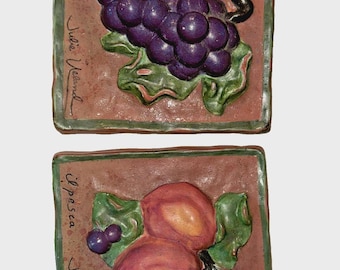 May include: Two square ceramic tiles with raised relief fruit designs. One tile features a cluster of purple grapes with green leaves, the other shows peaches. The tiles have a brown background and green borders.