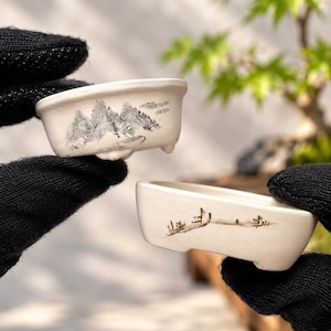 May include: Two small, off-white ceramic bonsai pots with short feet. The top pot features a detailed black ink-style mountain landscape painting. The bottom pot displays a minimalist black ink-style depiction of sparse trees and a horizon line.