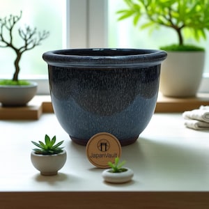 以下が含まれることがあります： まだら模様の釉薬を施した濃い青色の陶器鉢。丸い鉢は広い縁と茶色の底が特徴です。白い鉢に入った小さな多肉植物と木製のJapanVaultのロゴが鉢の前にあります。背景には盆栽があります。