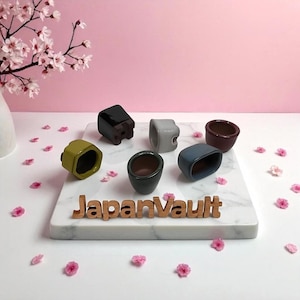 May include: A selection of small, colourful ceramic pots in various shapes and hues, including green, grey, and burgundy. The pots are arranged on a white marble surface with the text "JapanVault" in wooden letters. Pink flowers and a pink background complete the scene.