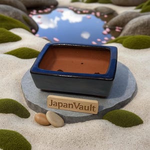 以下が含まれることがあります： 濃い青色の釉薬が施された長方形の盆栽鉢。内側は茶色。灰色の石の上に置かれ、近くには「JapanVault」と書かれた木の看板がある。背景には池と苔のあるミニチュアの禅庭園が写っている。